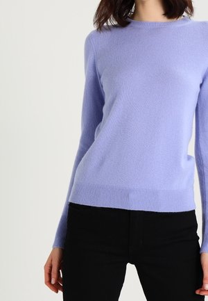 Žena v dlouhém levandulovém svetru s kulatým výstřihem a černých kalhotách, stojící před jednoduchým světlým pozadím.