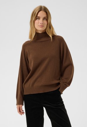 Personne aux cheveux blonds portant un pull à col roulé marron et un pantalon noir, debout devant un fond clair uni.