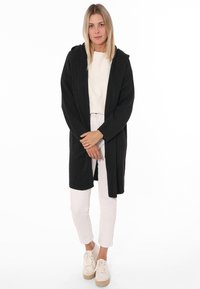 Schwarzer nahtloser langer Cardigan mit Kapuze, lockere Passform, strukturiertes Material, kombiniert mit einem hellen Oberteil und weißen Hosen. Weiße Schuhe runden das Outfit ab.