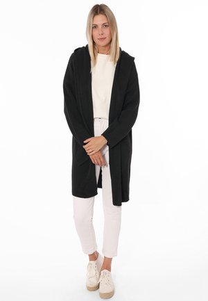 Czarny, dziergany kardigan z kapturem i otwartym przodem, z długimi rękawami i dwoma przednimi kieszeniami. Noszony na jasnym topie i białych spodniach.