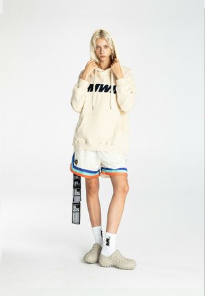 Persona joven con cabello rubio lleva una sudadera con capucha color crema, pantalones cortos deportivos blancos con rayas de colores, calcetines blancos y zapatos beige texturizados sin cordones.