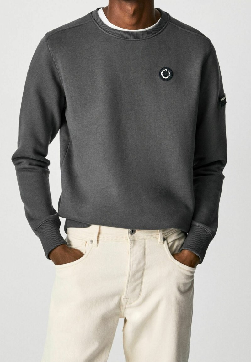 Homme portant un sweat-shirt à manches longues gris foncé avec un logo rond sur la poitrine et un pantalon beige clair, les mains dans les poches, sur un fond uni.