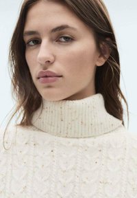 Kremni pulover s turtleneck ovratnikom, ki se ponaša z teksturirano pletenico in speckles po celotni površini ter rebrastim ovratnikom.