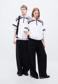 Λευκά πουκάμισα με φερμουάρ στον λαιμό, με αποχρώσεις μέντας και μαρούνης, συνδυασμένα με μαύρο παντελόνι φαρδύ. Οι λεπτομέρειες με σχέδια στους ώμους προσθέτουν υφή.