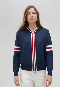 Marinblå zip-up hoodie med vit och röd randdesign längs framsidan och ärmarna, med en texturerad yta och en åtsittande passform.