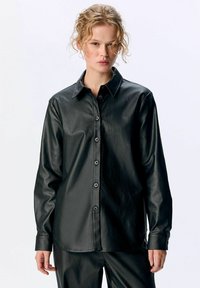 Chemise noire en simili cuir à boutons, avec un col, des manches longues et une coupe droite, présentant une texture lisse et cinq boutons à l'avant.