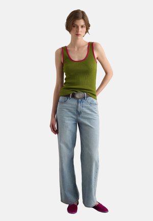 Femme portant un débardeur vert olive avec bordure rose, un jean large bleu clair avec ceinture, et des chaussures violettes à enfiler, debout sur fond blanc.