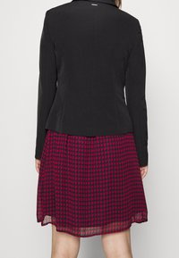 Černý blazer se strukturovaným designem, doplněný o šaty do kolen s červeno-černým kostkovaným vzorem. Hladká textilie.