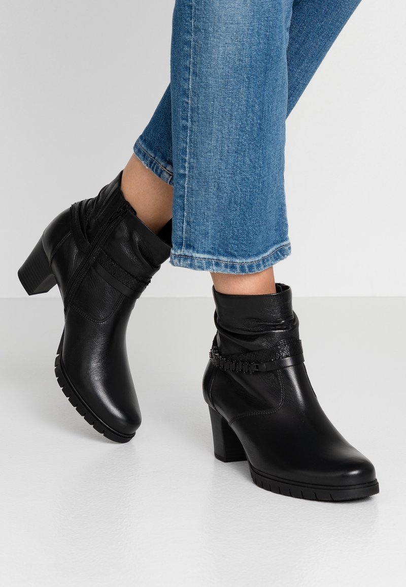 Bottes en cuir noires mi-mollet avec talons carrés, fermeture éclair latérale et bride décorative, portées avec un jean court en denim bleu sur un fond uni.