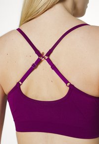 Sutiã roxo com alças cruzadas ajustáveis, apresentando ferragens metálicas nos pontos de conexão e uma textura de tecido suave e elástica.