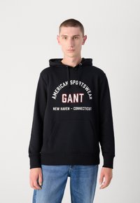 Čierna mikina s kapucňou a klokankou, s bielym a červeným textom: "AMERICAN SPORTSWEAR GANT NEW HAVEN - CONNECTICUT."