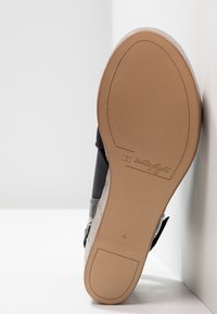 Semelle d'une sandale compensée noire inclinée contre un mur, montrant une semelle lisse couleur fauve avec le logo « butterfly light » et l'empreinte de taille 4.