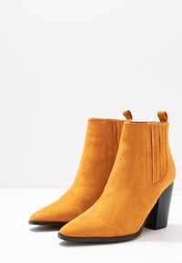 Bottes chevilles en daim jaune moutarde vif avec bouts pointus et panneaux élastiques latéraux ; talons empilés noirs et languettes de tirage en boucle.