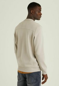 Felpa beige chiaro con polsini e orlo a costine, caratterizzata da un dettaglio di cucitura lungo la schiena. Indossata sopra una maglietta arancione a contrasto, abbinata a jeans scuri.