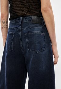 Tmavomodré rifľové nohavice majú voľný strih, dve zadné vrecká a čiernu koženú nášivku s logom na páse. Textúra je hladká.