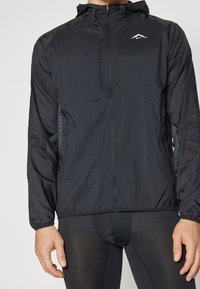 Lekka czarna kurtka do biegania z zamkiem z przodu, kapturem i elastycznymi mankietami. Posiada oddychające boczne panele z siateczki oraz małe logo na klatce piersiowej.