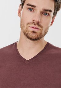 Bordowa koszulka z dekoltem w serek, wykonana z miękkiej bawełny. Dopasowany fason z krótkimi rękawami i delikatnymi detalami w przeszyciach na kołnierzyku oraz u dołu.