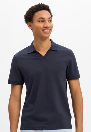Junger Mann, der lächelt und nach rechts schaut, trägt ein dunkelblaues kurzärmeliges Poloshirt mit kleinem gesticktem Logo auf der Brust.