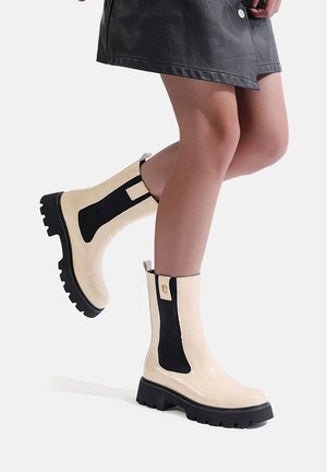 Stivali beige alla caviglia con pannelli laterali in elastico nero, finitura lucida, suola in gomma nera spessa e dettagli in bottone dorato sul retro.