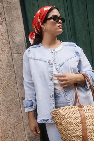 Chaqueta de mezclilla azul claro con botones plateados, camiseta blanca y bolsa de paja trenzada con asas de cuero marrón, complementada con una bufanda de estampado rojo.