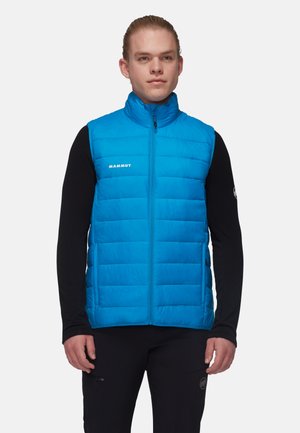 Junger Mann trägt eine leuchtend blaue gesteppte ärmellose Weste über einem schwarzen Langarmshirt und steht vor einem einfarbigen Hintergrund.