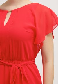 Vestido rojo de tela ligera, con mangas acampanadas, escote con abertura y una cintura ajustada con un detalle de cinturón atado.