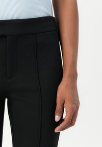 Gros plan d'une personne portant un pantalon noir ajusté avec un haut côtelé gris clair, montrant la taille, la hanche et la main droite reposant sur le côté.