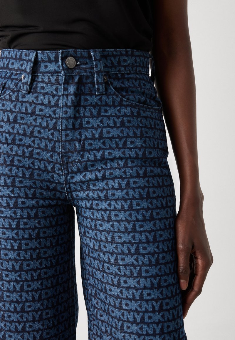 Jeans de mezclilla en color azul oscuro con un patrón repetido de logo "DKNY" en azul claro, cintura de altura media y silueta ajustada.