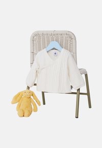 Krémový pletený sveter s výstrihom do V a káblovým vzorom, zobrazený na modrom vešiaku vedľa žltej plyšovej zajace, na bielom pozadí.