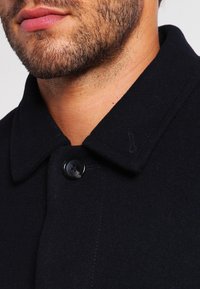 Cappotto di lana scuro con colletto a punta, dotato di chiusura a un bottone e dettagli di cucitura sottili che enfatizzano una texture morbida.