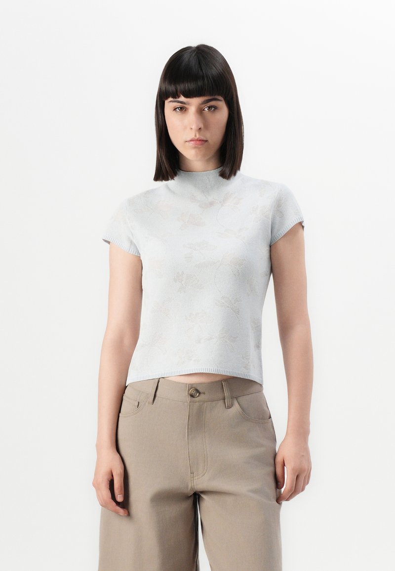 Lyseblå strikket topp med korterm, høyt krage, med et subtilt blomsterdesign, kombinert med beige høy-midje bukser.