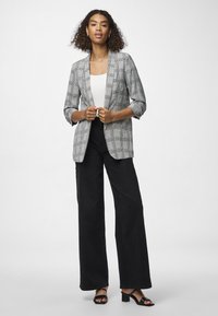 Blazer xadrez cinza com gola cachecol, mangas dobradas e tecido leve; combinado com calças pretas de perna larga e cintura alta e sandálias pretas de salto bloco.