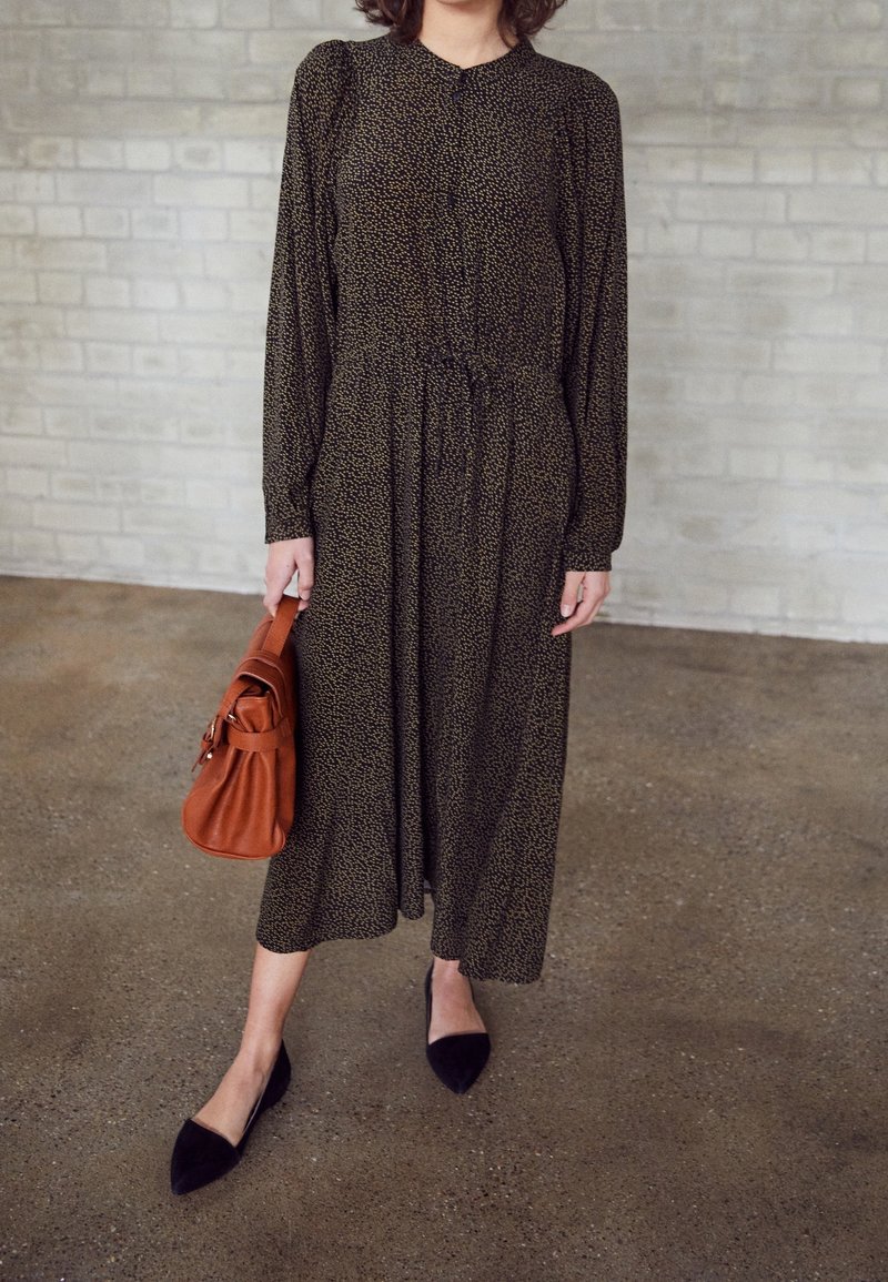 Robe longue à motifs noir et or avec une taille froncée, des manches longues et un col rond. Accessoirisée avec un sac à main marron et des ballerines noires.