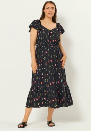 Femme portant une robe midi noire à petit imprimé floral rouge, blanc et vert, manches courtes bouffantes, et sandales plates noires, debout contre un mur blanc.