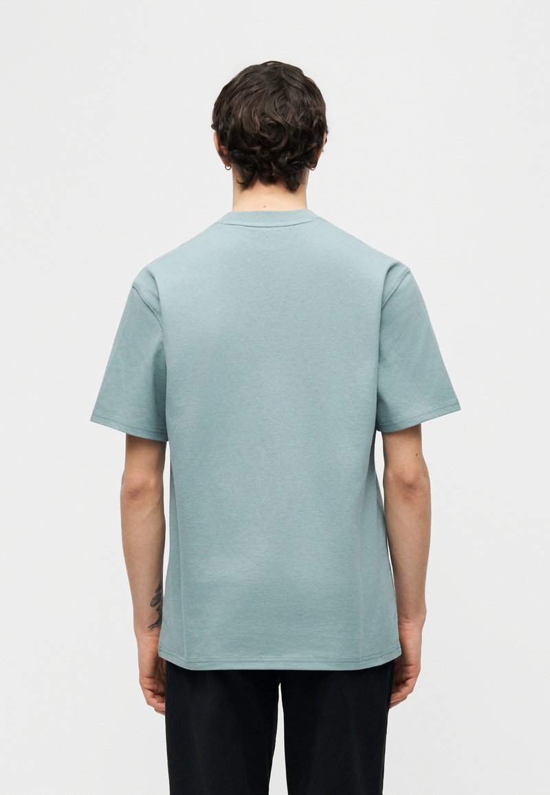 Uomo con capelli ricci corti che indossa una camicia celeste a maniche corte e pantaloni neri, in piedi di schiena contro uno sfondo semplice.