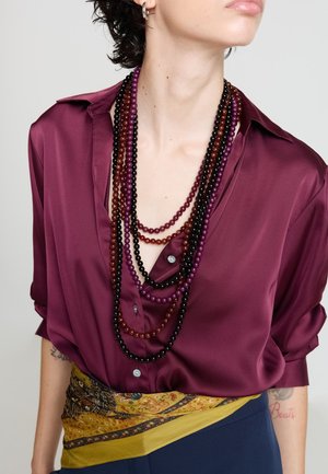 Samtige Bluse in Burgunderrot mit einem tiefen V-Ausschnitt, verziert mit mehreren geschichteten Perlenketten in Purpur-, Schwarz- und Bernsteintönen, ergänzt durch einen gemusterten Taillengürtel.