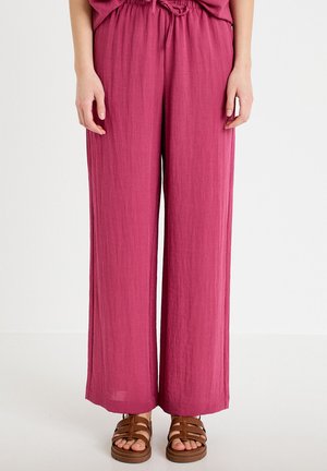 Person iført løstsiddende, højttaljede magentarøde bukser med bindebånd i taljen og brune sandaler med stropper, stående på et hvidt gulv.