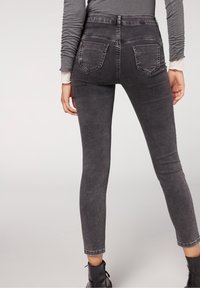 Persona vestida con jeans negros ajustados y desgastados, y botines negros, mostrada desde la cintura hacia abajo con las manos relajadas a los lados.