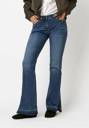 Flared, dark blue denim jeans with a high waist, visible stitching, and side slits at the hem. Paired with black pointed shoes.