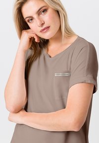 Bruine blouse met korte mouwen, een ronde halslijn en een klein zakje met een zilveren ritsaccent. Zachte stofstructuur, casual pasvorm.