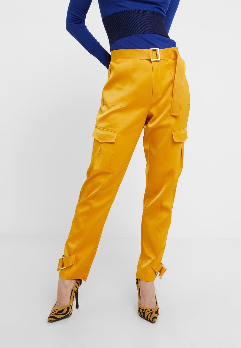 Femme portant un pantalon jaune vif avec des poches cargo et des détails à boucle, associé à des escarpins à rayures tigre et un haut bleu.