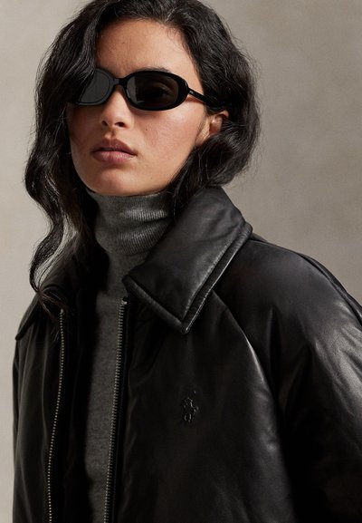 Jachetă bomber din piele neagră cu guler larg, purtată peste un pulover gri cu gât înalt. Ochelari de soare negri, de formă ovală, completează ținuta.