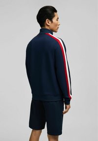 Marineblaues Sportjackett mit hohem Kragen, das rote und weiße Streifen an den Ärmeln aufweist. Getragen mit passenden marineblauen Shorts, die eine glatte Textur zeigen.