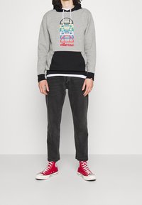 Felpa grigia con dettagli neri, grafica colorata sovrapposta sul davanti e logo rosso "ellesse". Abbinata a jeans neri e sneakers alta rosse.