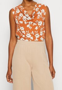 Ärmlös topp i rost med vitt blommönster, med en draperad halsringning. Kombineras med högmidjade, ljusbeiga, löst sittande byxor.