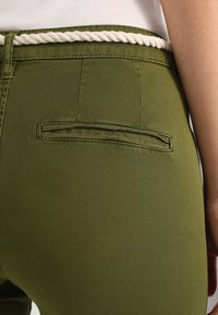 Olivgröna byxor har en slät struktur, en åtsittande design och en vävd repdetalj i midjan. En enkel bakficka är synlig.