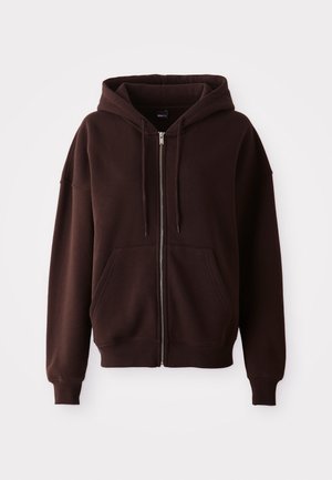 Ruda zip-up džemperis, pagamintas iš minkštos medžiagos, turi didelę kapučiną, priekinės kišenes ir gomurius bei apačią su gumbais. Nėra matomų raštų ar akcentų.