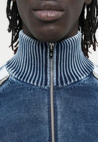 Πουλόβερ με φερμουάρ και ψηλό λαιμό σε βαθμιαίο μπλε πλεκτό, με ριμπ υφή και αντίθετες ανοιχτόχρωμες μπλε λεπτομέρειες στους ώμους.