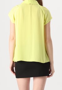 Blusa gialla a maniche corte con colletto, realizzata in tessuto leggero. Presenta un orlo curvo e un look sovrapposto su una mini gonna nera.