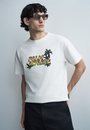 Νέος άνδρας με σγουρά σκούρα μαλλιά, φοράει γυαλιά ηλίου, λευκό T-shirt με γράμματα "PALM ANGELS" και σχέδιο φοίνικα, καθώς και μαύρο παντελόνι, στέκεται μπροστά σε φωτεινό τοίχο.